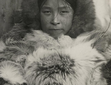 Black and white photo of an Inuit woman called Aqattaq. Aqattaq stares forward with a nutural expression wearing a fur top and fur hood.