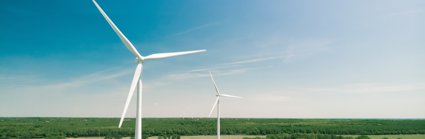 A view of two white wind turbines, located in flat countryside with fields and forest