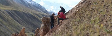 Two researchers sit on a sloping valley side while one stands, facing the others. They are all writing in notebooks.