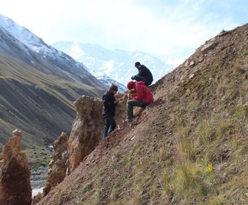 Two researchers sit on a sloping valley side while one stands, facing the others. They are all writing in notebooks.