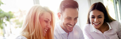 Three people sitting at a table looking at screens whilst smiling.