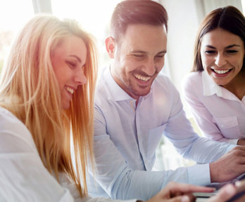 Three people sitting at a table looking at screens whilst smiling.