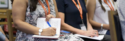 A person writing in a notebook while listening to a conference presentation, seated next to two other conference delegates