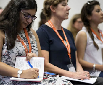 A person writing in a notebook while listening to a conference presentation, seated next to two other conference delegates