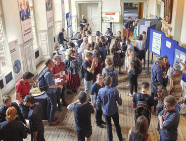 A group of people wearing lanyards in a stately Main Hall. The room features display boards and posters.