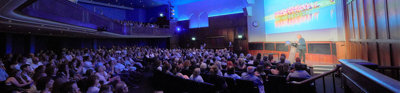 Audience members attending a lecture. One speaker is standing in front of a projection screen on a stage presenting.