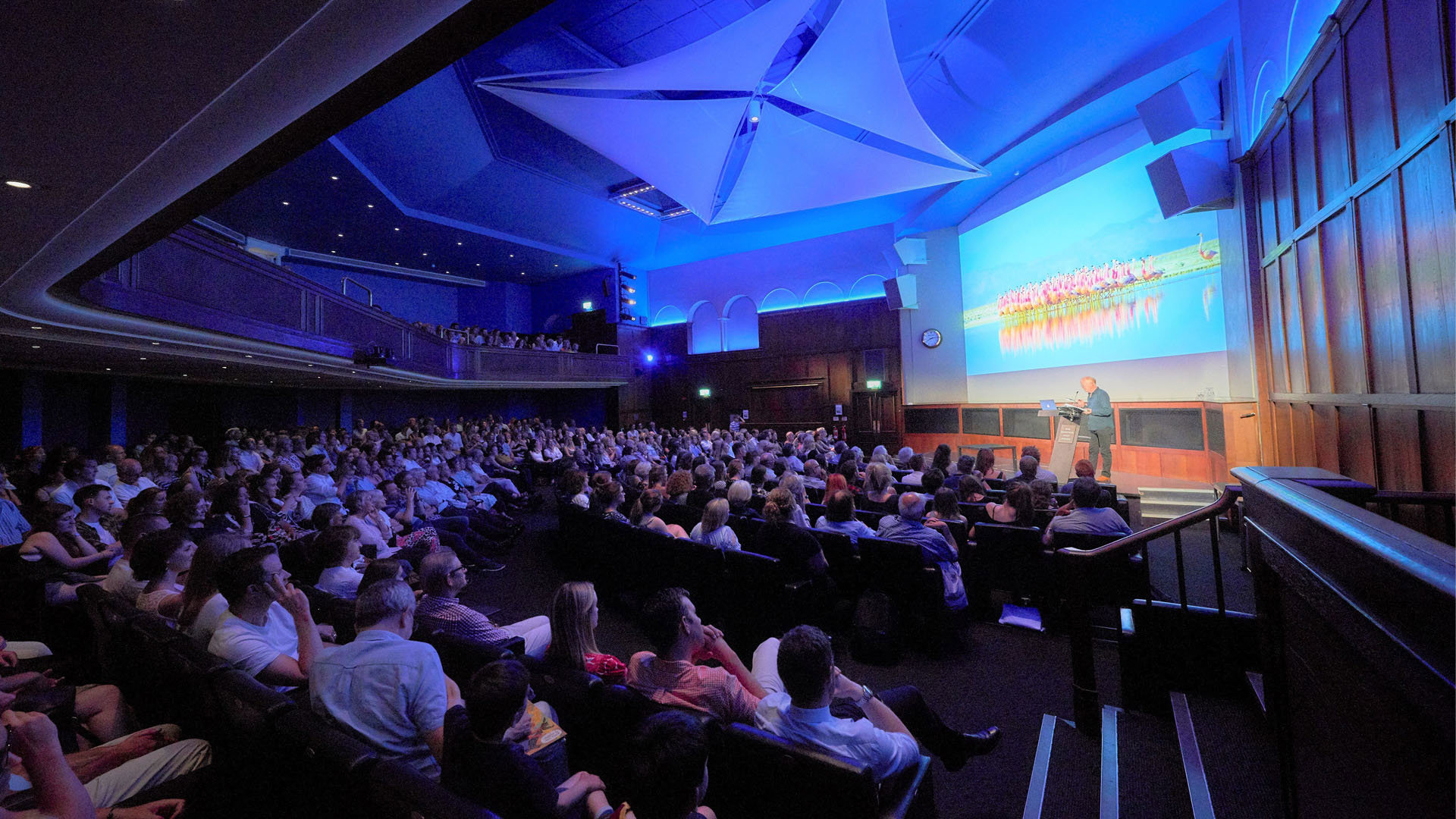 Audience members attending a lecture. One speaker is standing in front of a projection screen on a stage presenting.