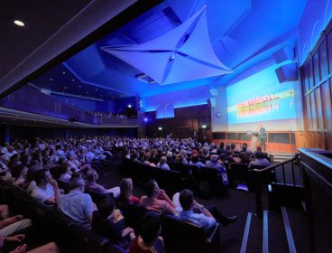 Audience members attending a lecture. One speaker is standing in front of a projection screen on a stage presenting.