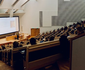 Lecture theatre with students sitting in staggered rows of seats. The rows are faced towards a stage with someone giving a presentation in front of a screen. 