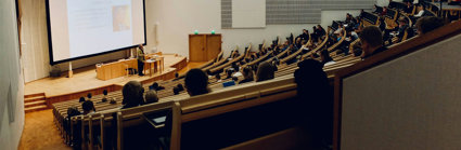 Lecture theatre with students sitting in staggered rows of seats. The rows are faced towards a stage with someone giving a presentation in front of a screen. 