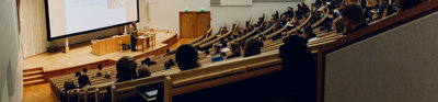 Lecture theatre with students sitting in staggered rows of seats. The rows are faced towards a stage with someone giving a presentation in front of a screen. 