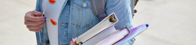 A student stands on a pavement holding two text books, a folder and notebook in one hand whilst holding on to their headphone cords with the other.