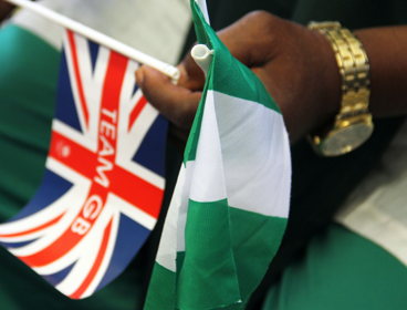 A person holds both the red, white and blue union jack alongside the white and grey Nigerian flag. Their shirt is white and toursers are green, matching the flag of Nigeria.