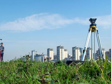 Surveying tool set up on a tripod in a field. There are skyscrapers in the background below a clear sky. 