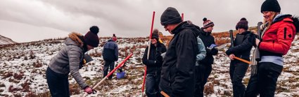 A group of 7 secondary school pupils holding ranging poles standing on a snowy hillside, they are smiling. 