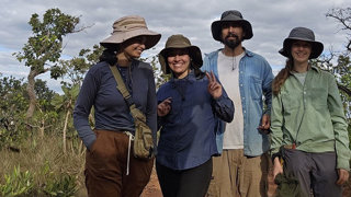 A group of four researchers posing for a group photo in a savanna landscape, on a dry brown dusty road. There is tall grass and small tress behind them.  