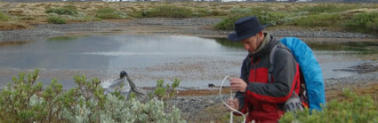 Person setting up a measuring device in front of a glacier and lake. The person is wearing a hat and rucksack.