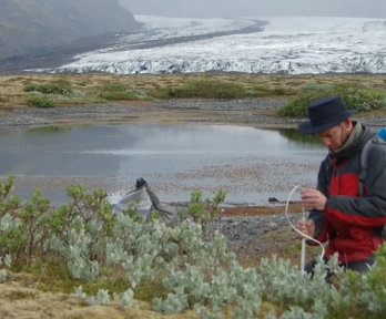 Person setting up a measuring device in front of a glacier and lake. The person is wearing a hat and rucksack.