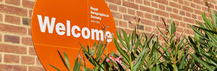 A circular orange sign reading 'Royal Geographical Society with IBG' and 'Welcome', next to a green plant with pink flowers.