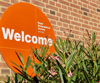 A circular orange sign reading 'Royal Geographical Society with IBG' and 'Welcome', next to a green plant with pink flowers.