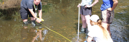 A grop of teachers are standing in a shallow river, two are holding a tape measure across the river to measure its width. Two are standing by and watching.