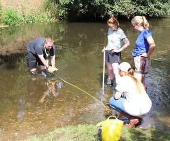 A grop of teachers are standing in a shallow river, two are holding a tape measure across the river to measure its width. Two are standing by and watching.