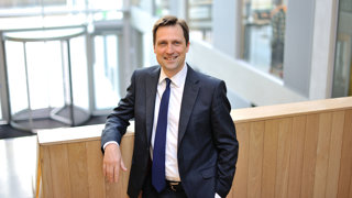 A man in suit and tie posing for a photo while leaning against a wooden handrail inside a bright office.
