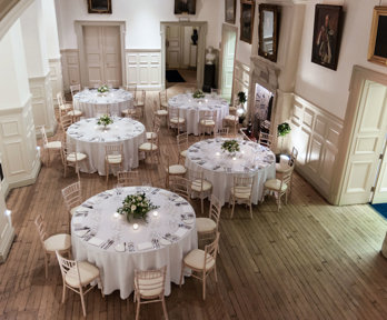 Five round tables with wooden chairs and white linen setup for a dinner in the Main Hall.