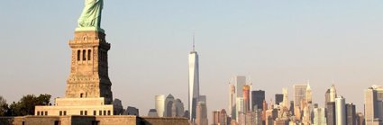 A view from the river of New York City, showing the Statue of Liberty and a skyscraper horizon. The sun is low in the sky so the light is soft