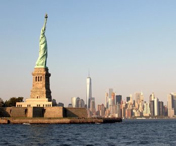 A view from the river of New York City, showing the Statue of Liberty and a skyscraper horizon. The sun is low in the sky so the light is soft
