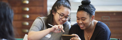 Two young people sat smiling while looking at a phone together. In the background are large woodend map drawers.