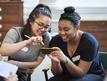 Two young people sat smiling while looking at a phone together. In the background are large woodend map drawers.