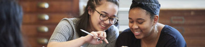 Two young people sat smiling while looking at a phone together. In the background are large woodend map drawers.