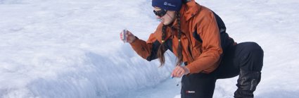 A researcher bending down by a crack in a glacier holding a transparent tube and looking at the contents.