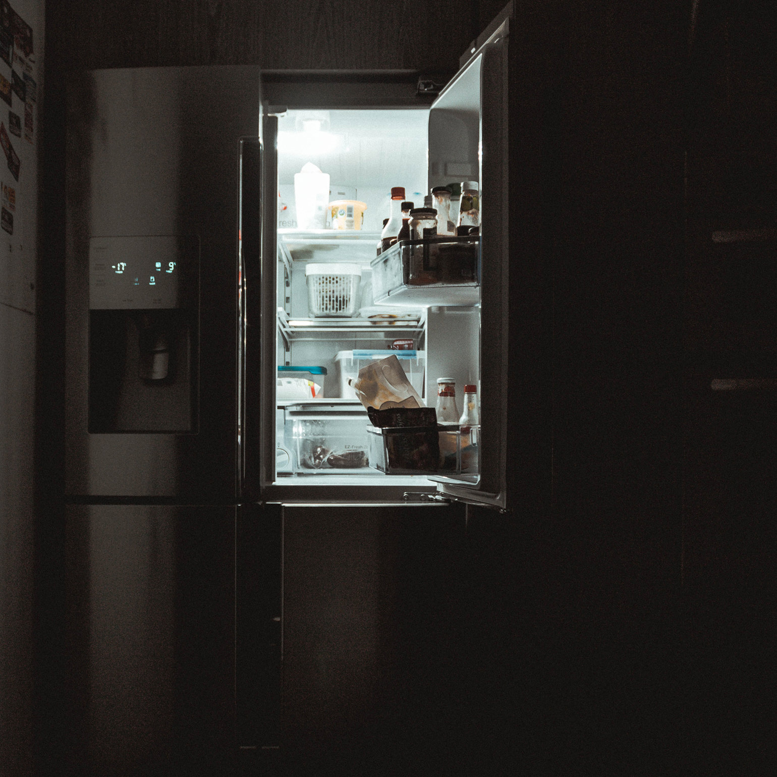 A dark room with an open fridge, so the light of the firdge is all you can see. The fridge has food and bottles in it