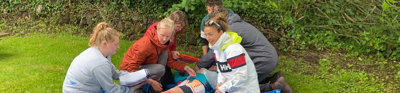 A group of people sitting on tarp in a field of grass, learning how to do first aid on a person that is laying and pretending to be injured.