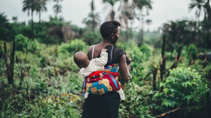 Woman farming in tropical surroundings carrying a child on her back hal wrapped in a colourful cloth.