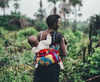 Woman farming in tropical surroundings carrying a child on her back hal wrapped in a colourful cloth.