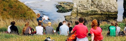 A group of young people sit on a seaside cliff, listening to a presenter in the front who's writing on a paper pad.