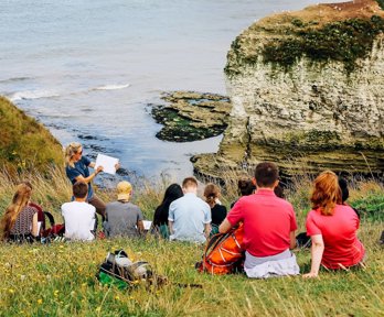 A group of young people sit on a seaside cliff, listening to a presenter in the front who's writing on a paper pad.