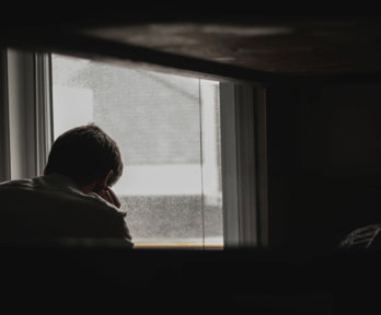Person faced away from the viewer looks outside of the window whilst their head rests on their hand. The room around them is dark and the landscape ouside the window is grey and blurred.