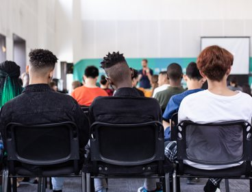 A view of a classroom from behind, with three students in the forefront of the picture shown from behind, watching the teacher