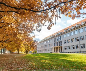 Large stately building fronted by green lawn and deciduous, golden and orange trees.