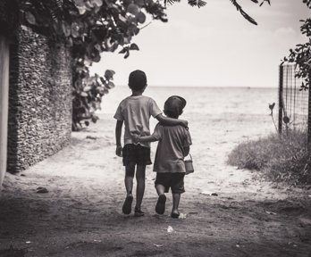 Two children with their arms around one another walking towards a beach