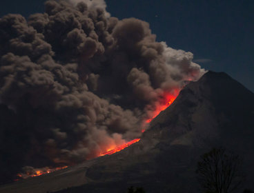 A volcano eruption with large smoke plumes rising into a dark sky and lava flowing down the side of the volcano.