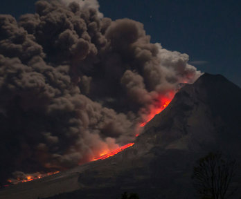 A volcano eruption with large smoke plumes rising into a dark sky and lava flowing down the side of the volcano.