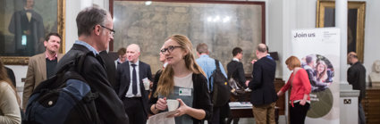 People stand scattered throughput the Royal Society map room. Most people appear to be mid conversation. Maps and portraits fill the walls behind.