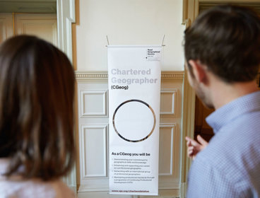 Two people at the Society look and point at a sign that has information about becoming a Chartered Geographer.