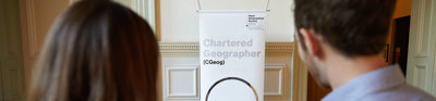 Two people at the Society look and point at a sign that has information about becoming a Chartered Geographer.