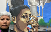 Laura Pulido taking a selfie in front of an artwork depicting a person using an asthma inhaler, a set of swings on a playground and a factory pipes with exhaust fumes.